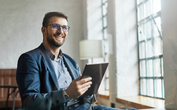 Términos Banca de inversión Foto de un hombre con barba y gafas de marco oscuros, vestido con camisa y saco. Tiene una tableta en sus manos conociendo información de los términos banca de inversión y mira hacia el horizonte sonriendo.